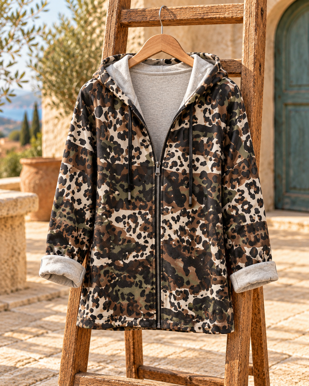 Bodegón de chaqueta de camuflaje con capucha y cremallera, en percha de madera sobre una terraza mediterránea.