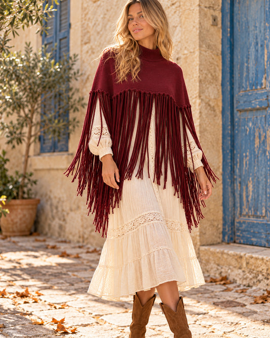Poncho corto de cuello alto con flecos largos en color burdeos, llevado sobre vestido marfil.