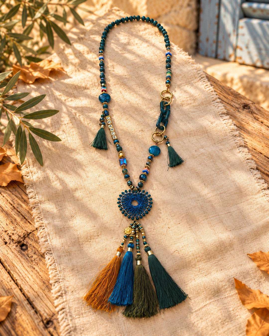 Collar largo boho medallón azul · borlas multicolor, bodegón sobre tejido claro