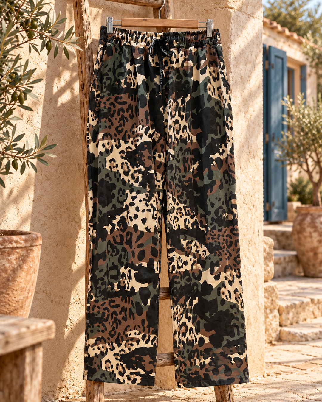 Bodegón del pantalón recto de camuflaje, completo y en percha sobre una escalera de madera en un patio mediterráneo.