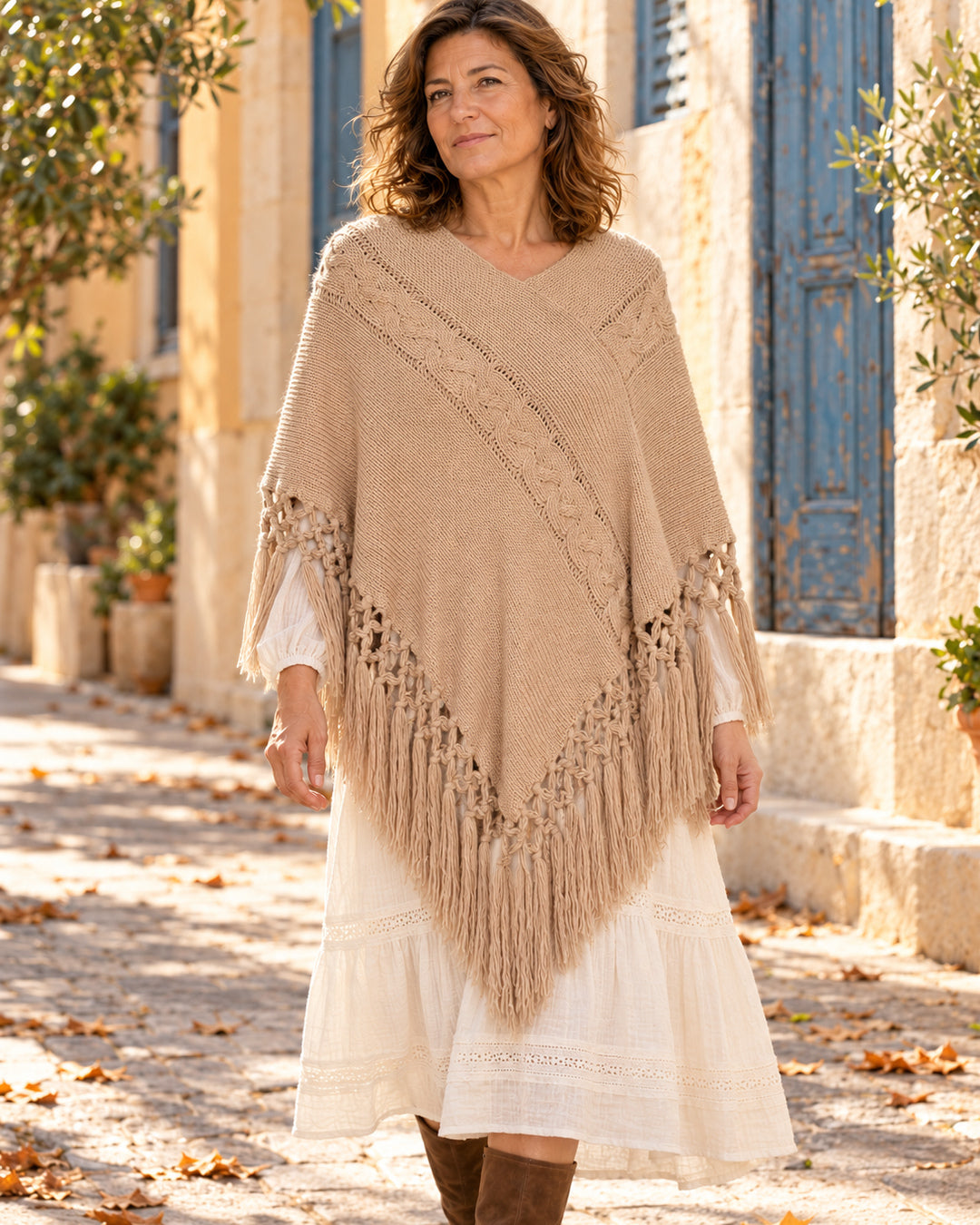 Poncho cerrado de punto con escote en V y flecos en color camel, llevado sobre vestido marfil.