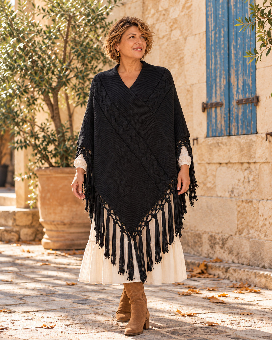 Poncho cerrado de punto con escote en V y flecos en color negro, llevado sobre vestido marfil.