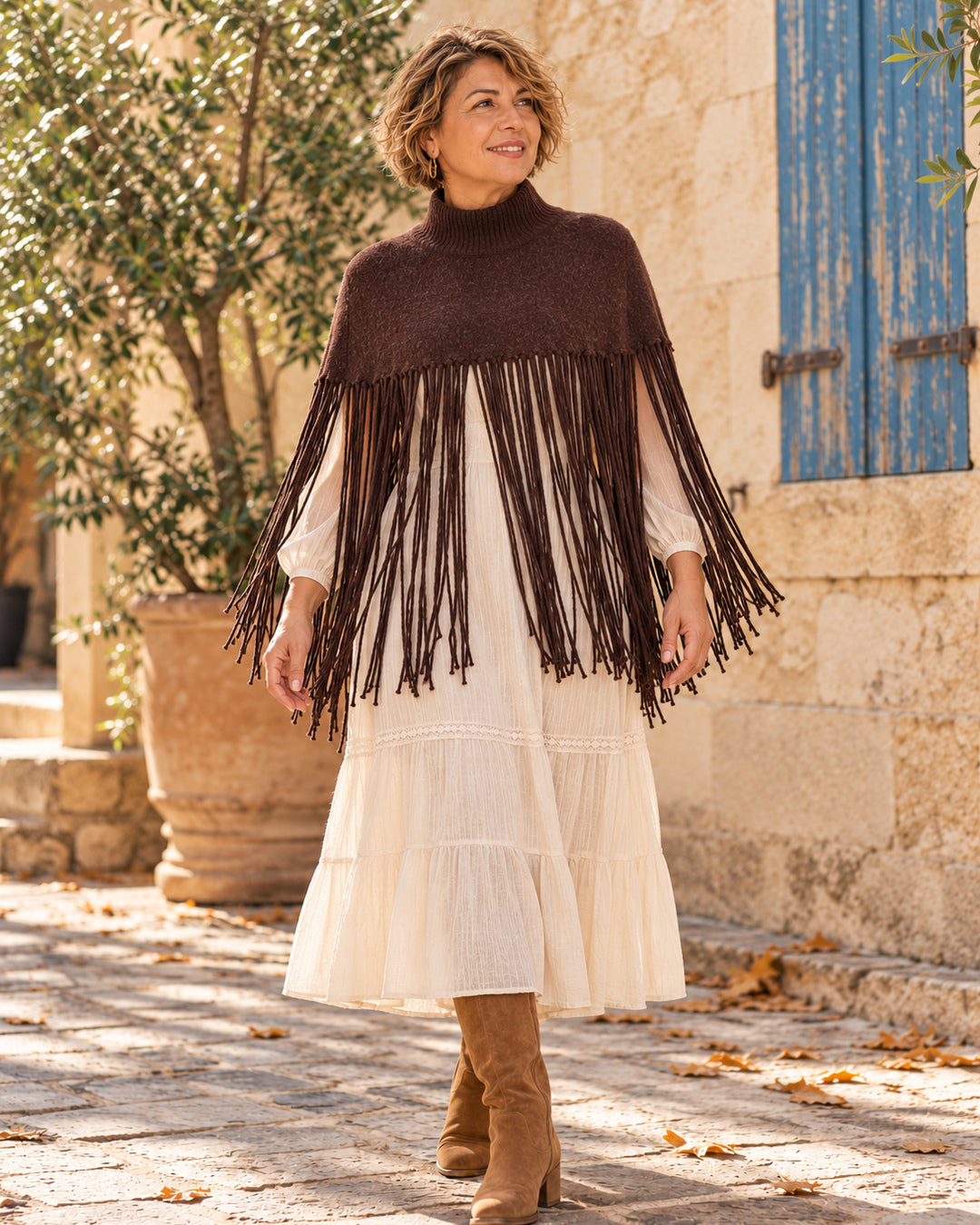 Poncho corto de cuello alto con flecos largos en color chocolate, llevado sobre vestido marfil.
