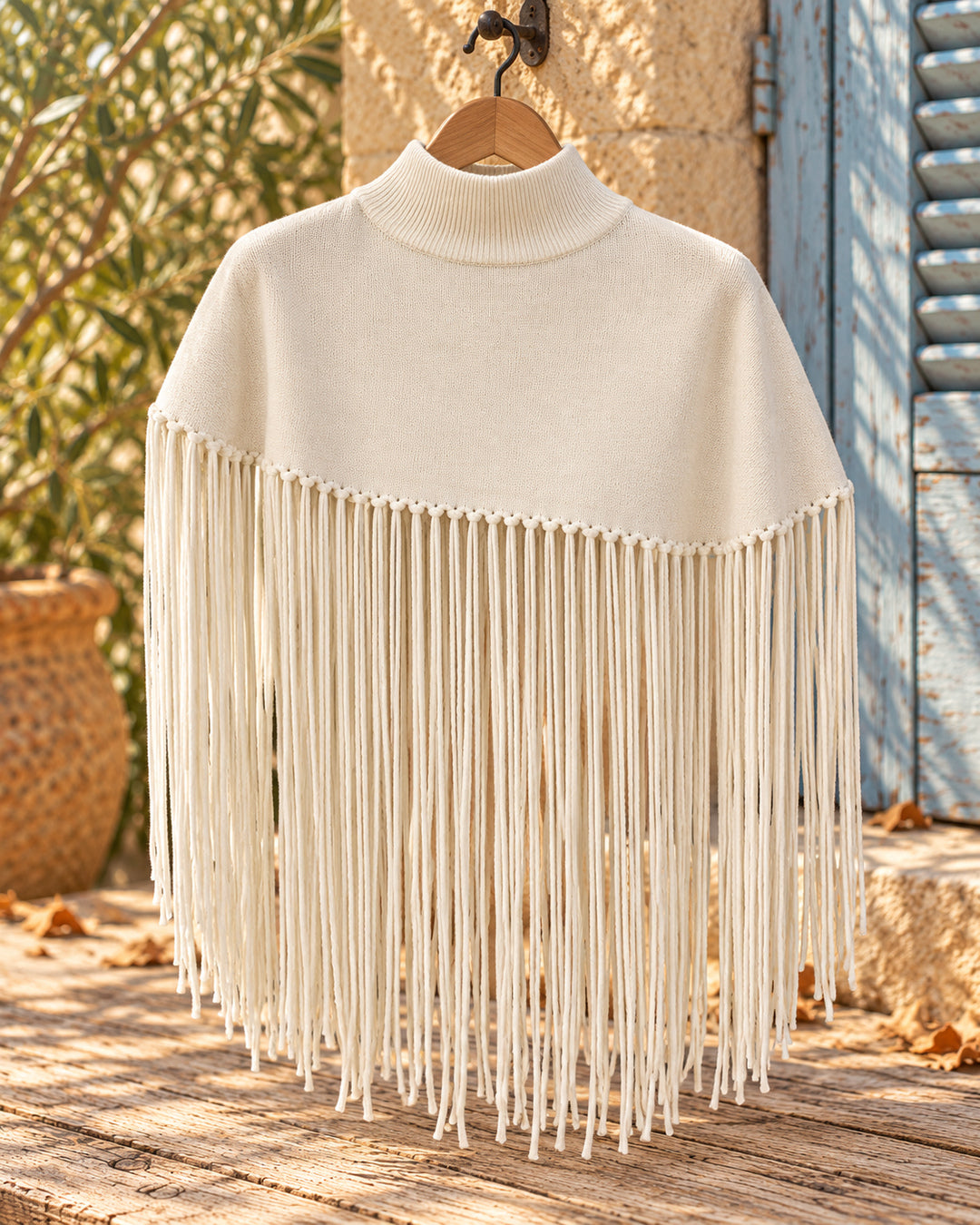 Poncho corto de cuello alto con flecos largos en color crema, vista del producto en percha.