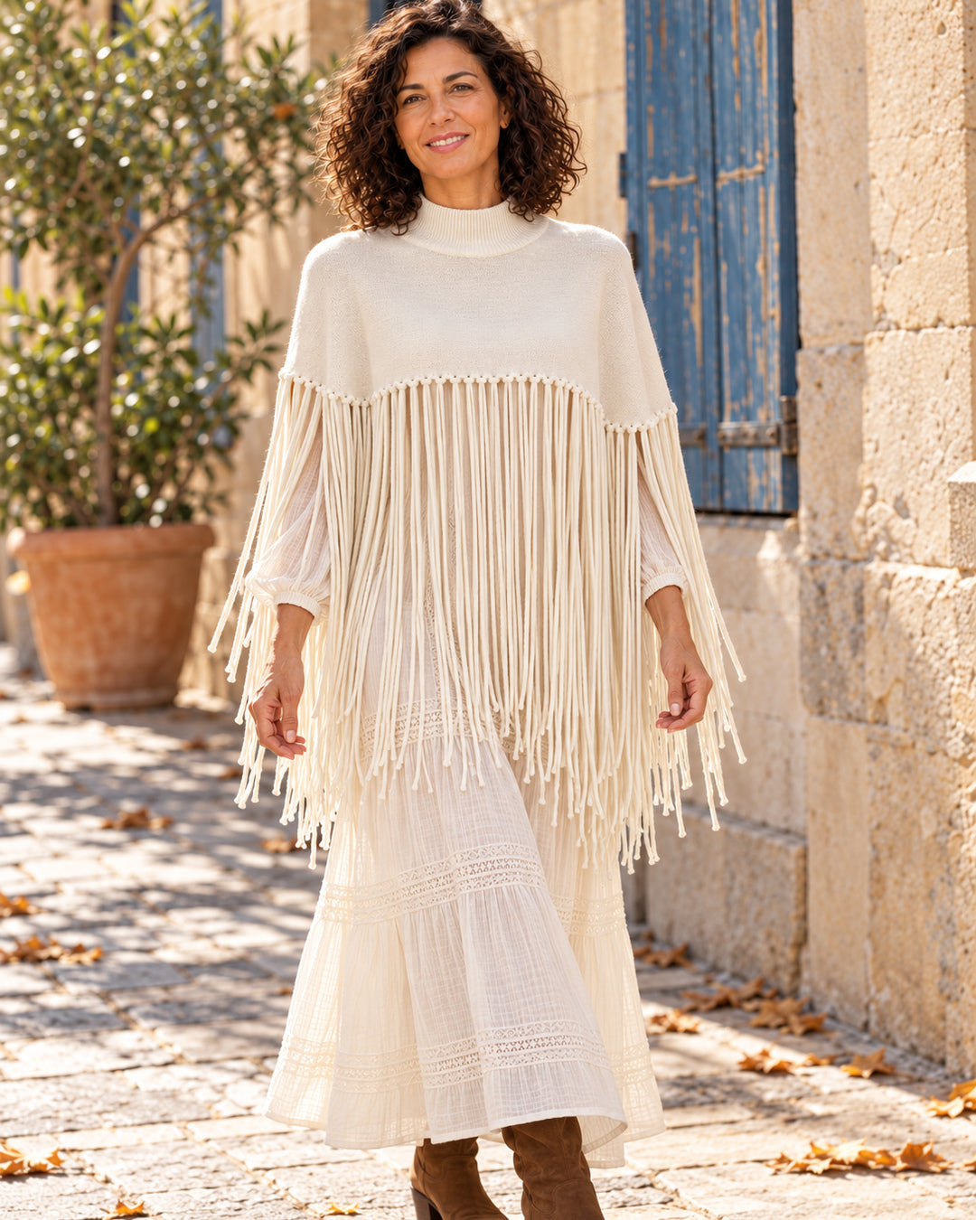 Poncho corto de cuello alto con flecos largos en color crema, llevado sobre vestido marfil.